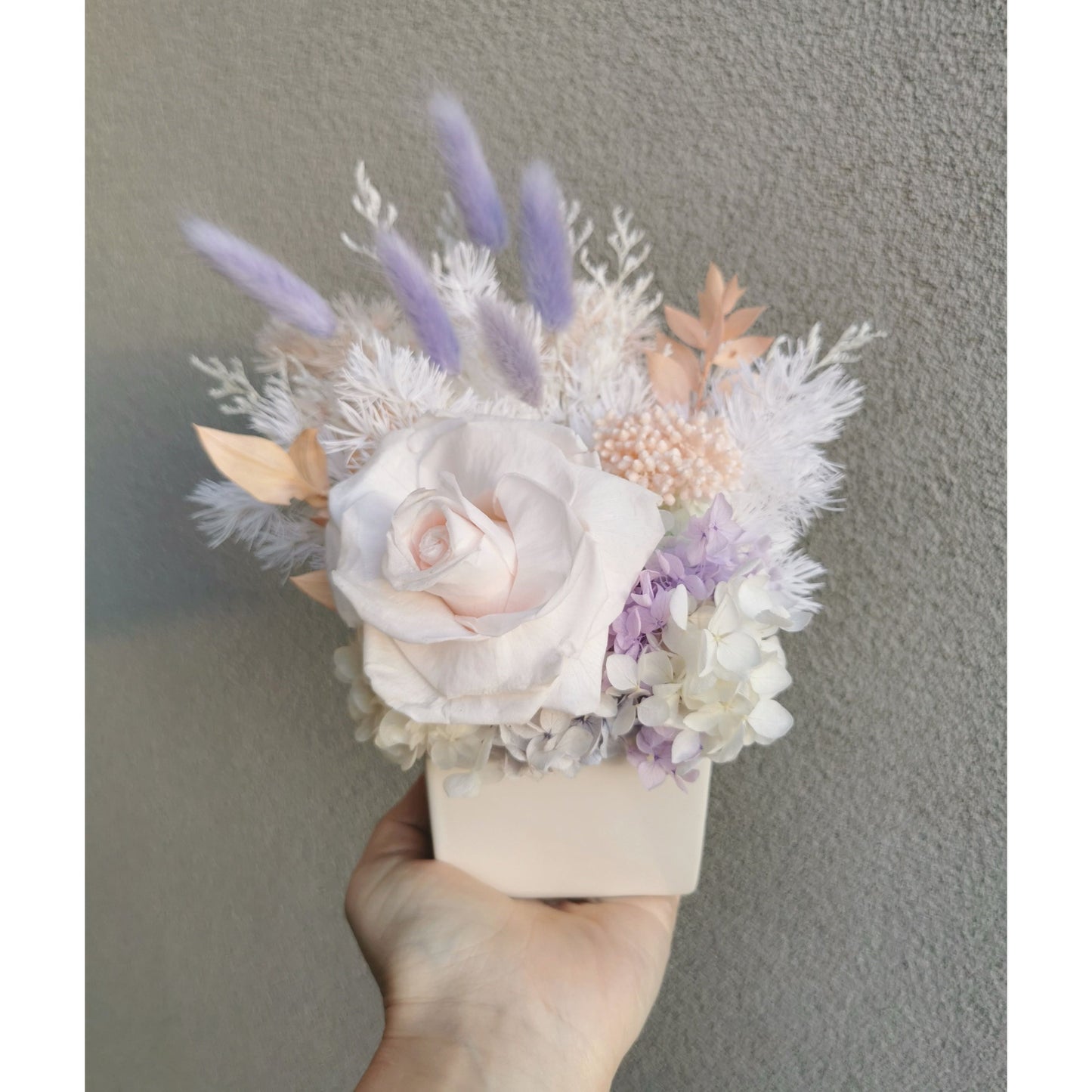 Pastel coloured dried & preserved flower arrangements in white cube pot. Features a soft pink preserved rose. Picture shows arrangement being held by hand against a blank wall.