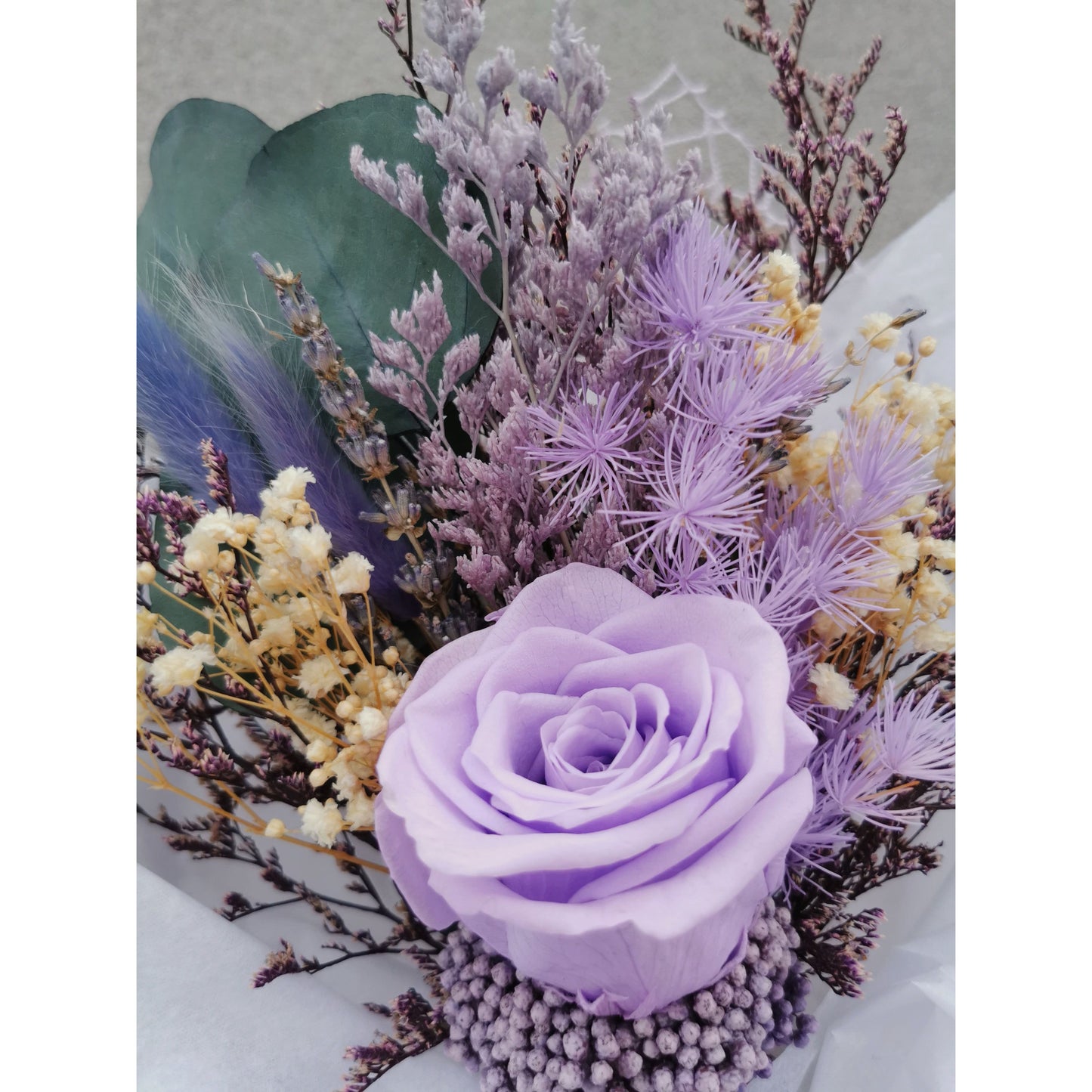 Small bunch of dried & preserved flowers in purple shades, green & cream and featuring a preserved purple rose. Wrapped ready for gifting. Pic shows bunch held up against a wall with a full close up of the flowers
