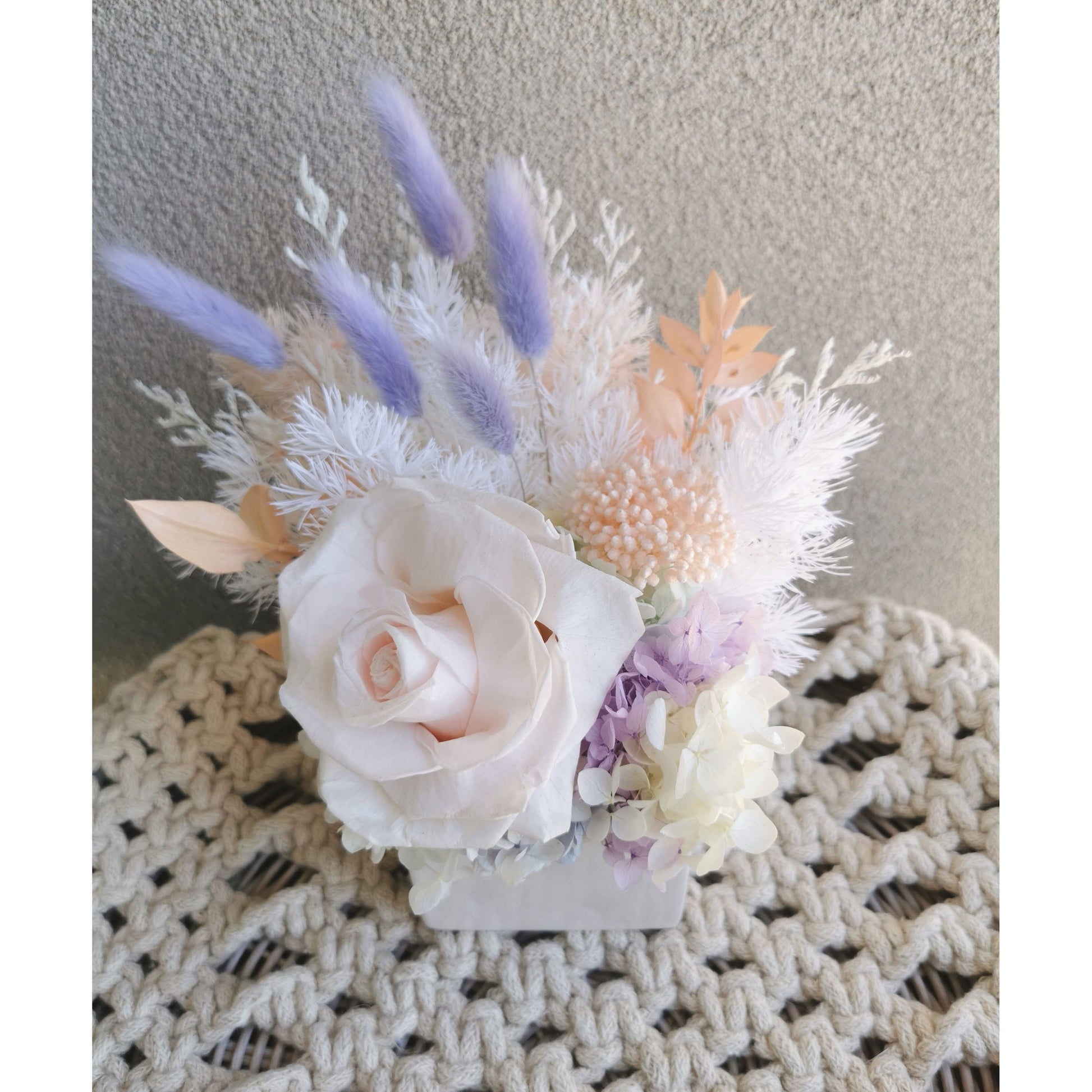 Pastel coloured dried & preserved flower arrangements in white cube pot. Features a soft pink preserved rose. Picture shows arrangement sitting on a table against a blank wall with a view of the flowers from above