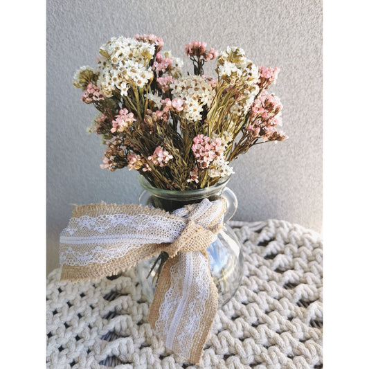 Dried pink & white oxeye daisy flowers in glass vase with lace hessian ribbon weaved through handles of vase. Picture shows arrangement sitting on table against a blank wall