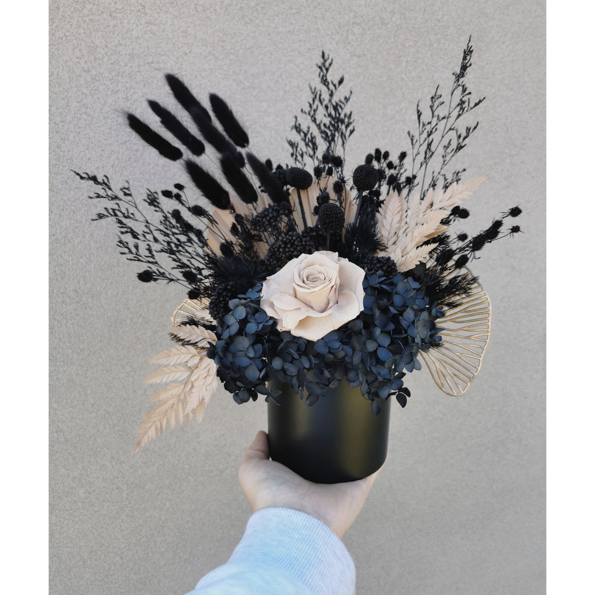 Black, nude & gold dried flowers set in to a black pot. Picture shows arrangement being held up against a blank wall