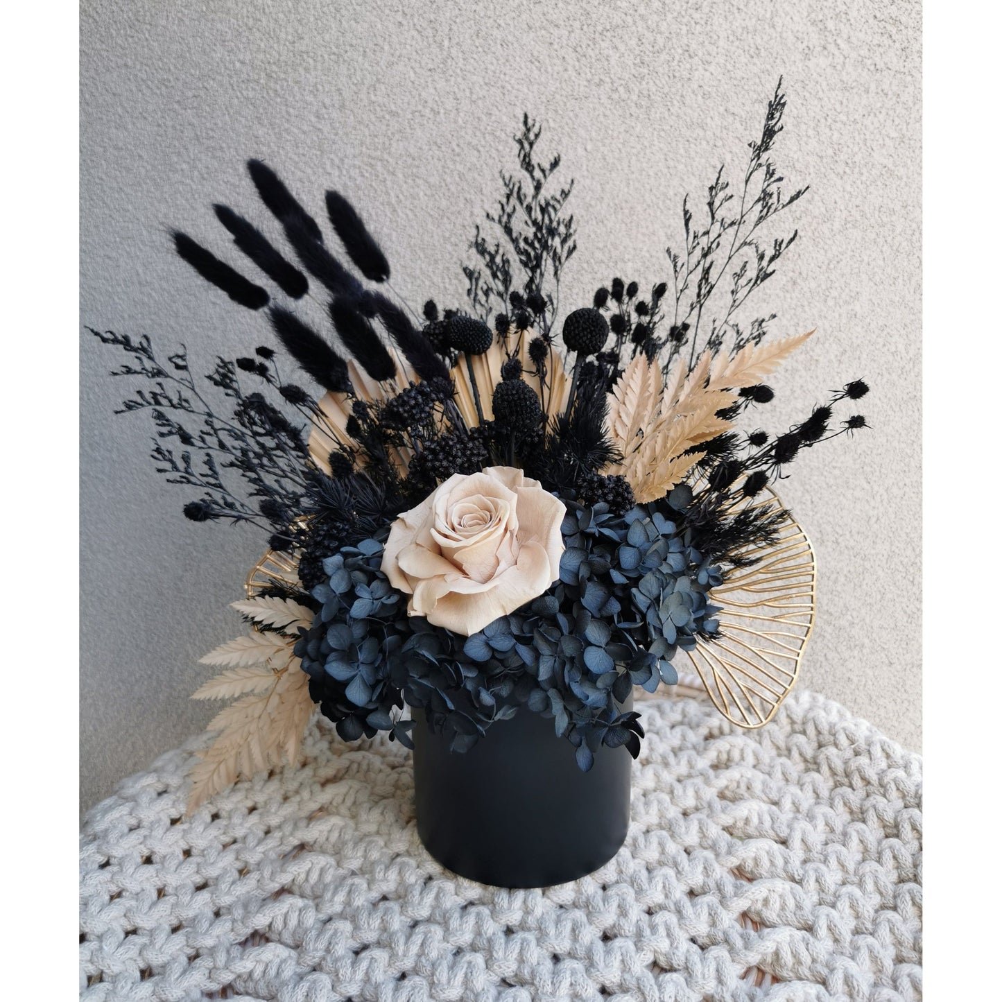 Black, nude & gold dried flowers set in to a black pot. Picture shows arrangement sitting on a tray being held up against a blank wall