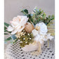 Dried, preserved and artificial flowers in colours green, white and neutral and set in to a rustic trough style pot. Picture is taken of the flower arrangement on a table against a blank wall on an angle