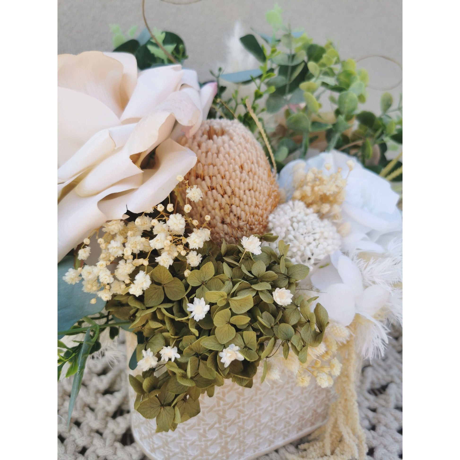 Dried, preserved and artificial flowers in colours green, white and neutral and set in to a rustic trough style pot. Picture is taken of the flower arrangement on a table against a blank wall with a close up view of the flowers