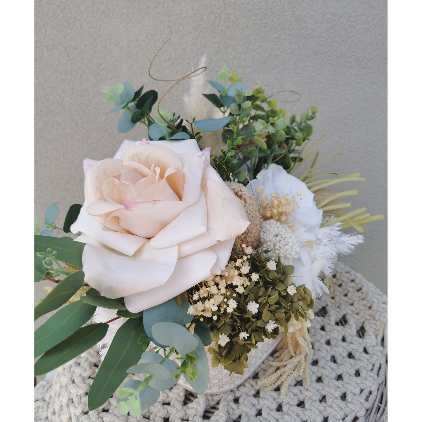 Dried, preserved and artificial flowers in colours green, white and neutral and set in to a rustic trough style pot. Picture is taken of the flower arrangement on a table against a blank wall on a side ono view angle