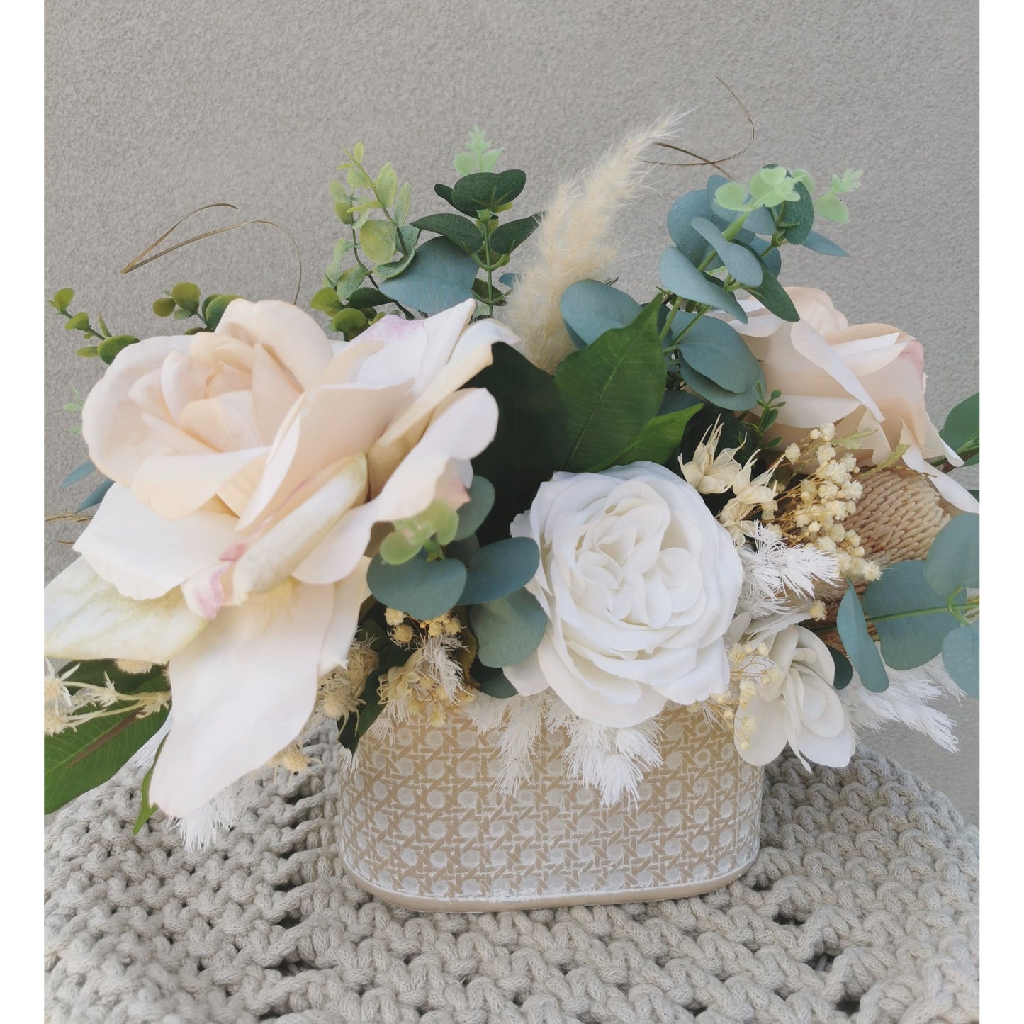 Dried, preserved and artificial flowers in colours green, white and neutral and set in to a rustic trough style pot. Picture is taken of the flower arrangement on a table against a blank wall showing the reverse side of the arrangement
