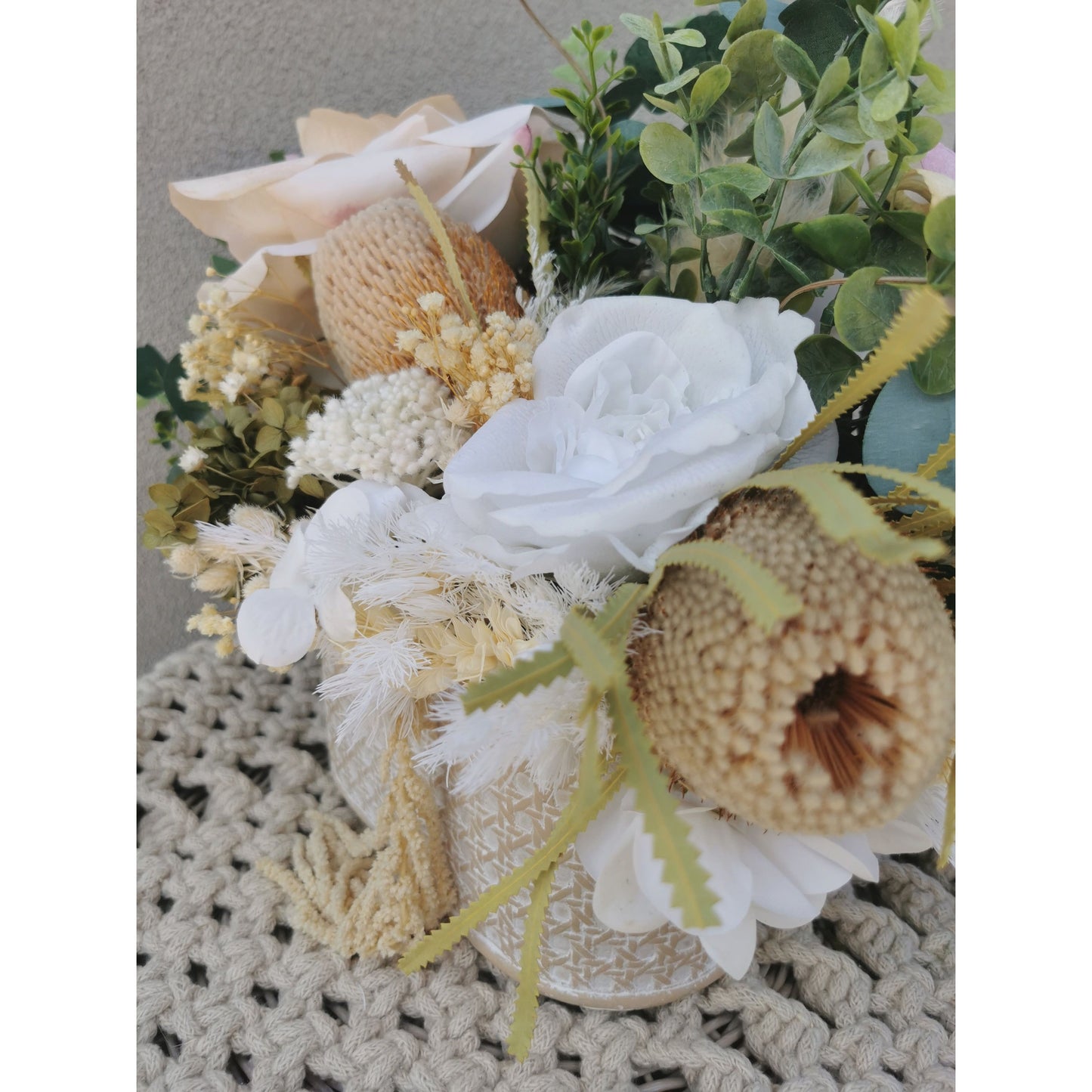 Dried, preserved and artificial flowers in colours green, white and neutral and set in to a rustic trough style pot. Picture is taken of the flower arrangement on a table against a blank wall on a side end angle view