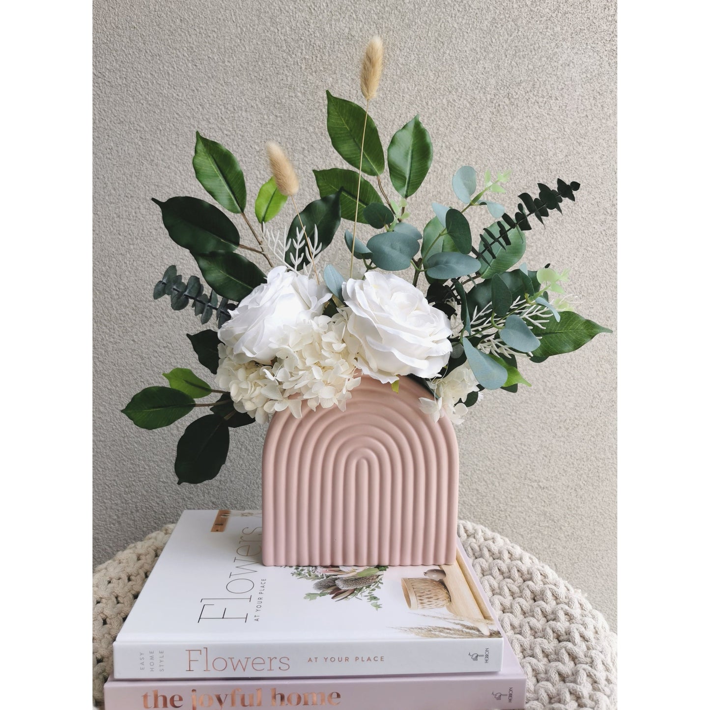 Green & white flowers consisting of dried, preserved and artificial flowers and set in to a pink arch vase. Picture shows the flower arrangement sitting on design books on a table against a blank wall