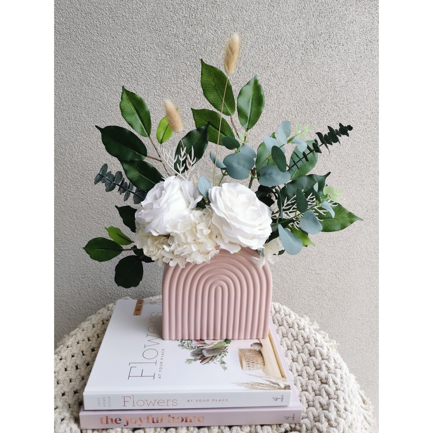 Green & white flowers consisting of dried, preserved and artificial flowers and set in to a pink arch vase. Picture shows the flower arrangement sitting on design books on a table against a blank wall with a view of the flowers from birds eye view