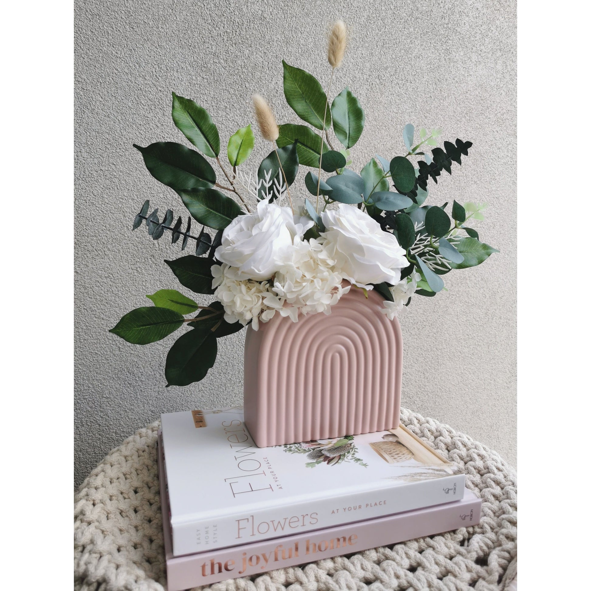 Green & white flowers consisting of dried, preserved and artificial flowers and set in to a pink arch vase. Picture shows the flower arrangement sitting on design books on a table against a blank wall and sitting on an angle to the right