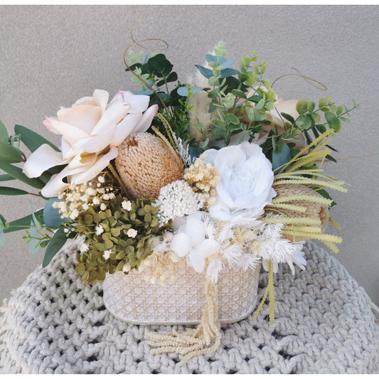Dried, preserved and artificial flowers in colours green, white and neutral and set in to a rustic trough style pot. Picture is taken of the flower arrangement on a table against a blank wall