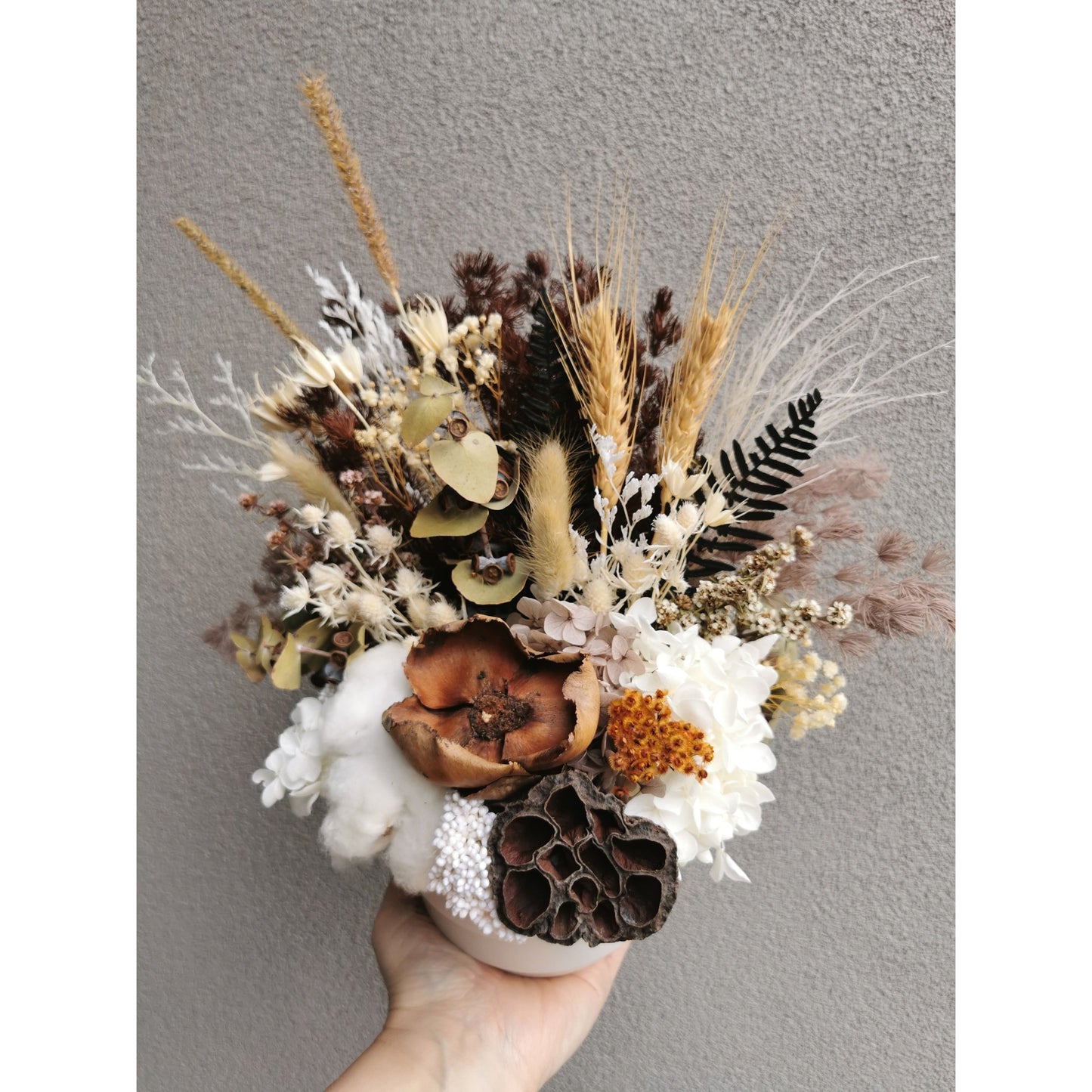 Dried & Preserved native flower arrangement in white pot. Picture shows flower arrangement being held up by hand against a blank wall and held at an angle