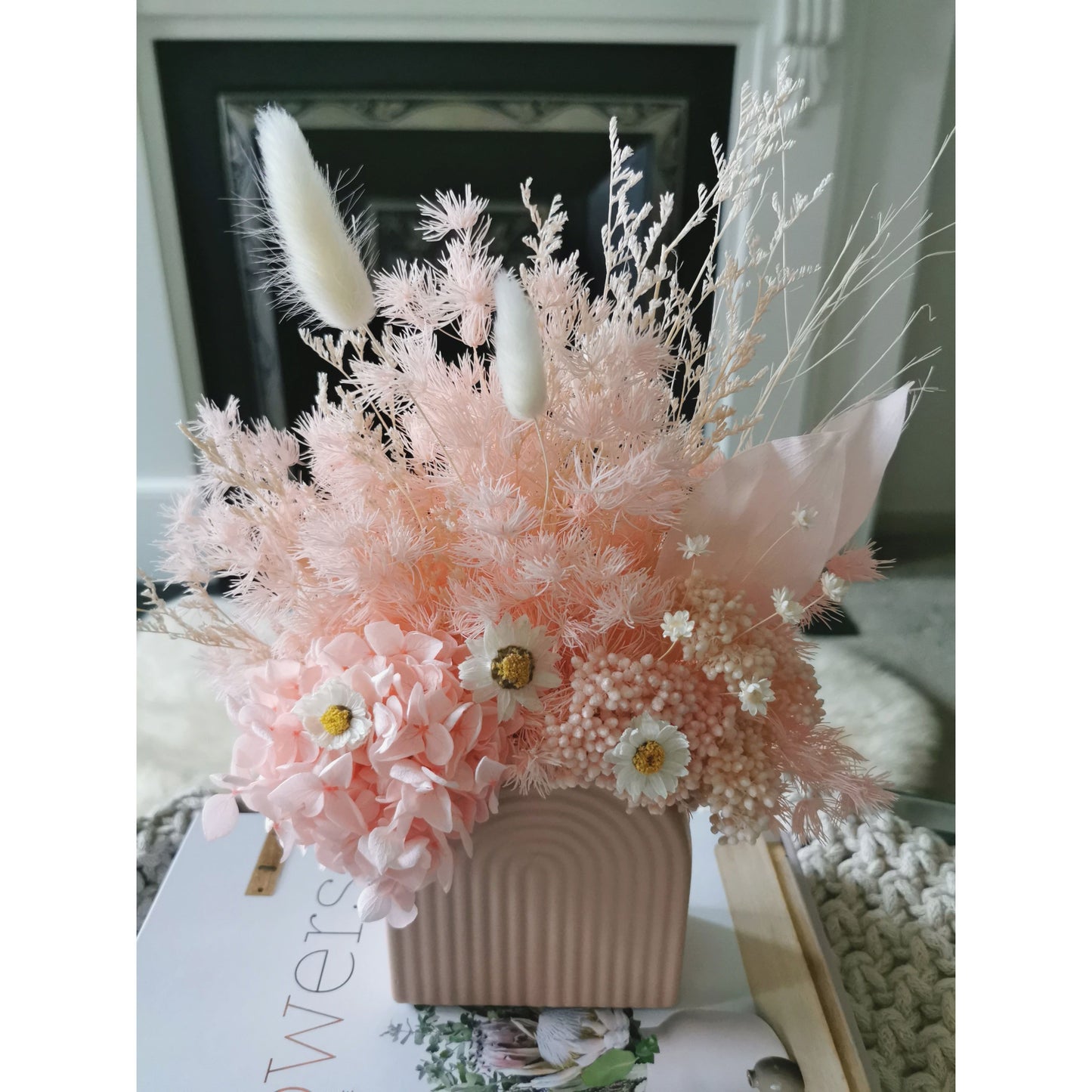 Dried & Preserved flower arrangement in pretty pink with 3 white daisies. Flowers are set in to a mini pink arch vase. Picture is showing the arrangement sitting on top of books on a table. Arrangement looking down from the top of the arrangement