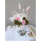 Dried & Preserved flower arrangement in pink, green & white featuring pink zinnia flowers. Photo shows arrangement sitting on books on a table against a blank wall