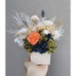 Dried & preserved flower arrangement with navy blue, green, white & orange flowers. Picture shows the arrangement being held by hand against a blank wall