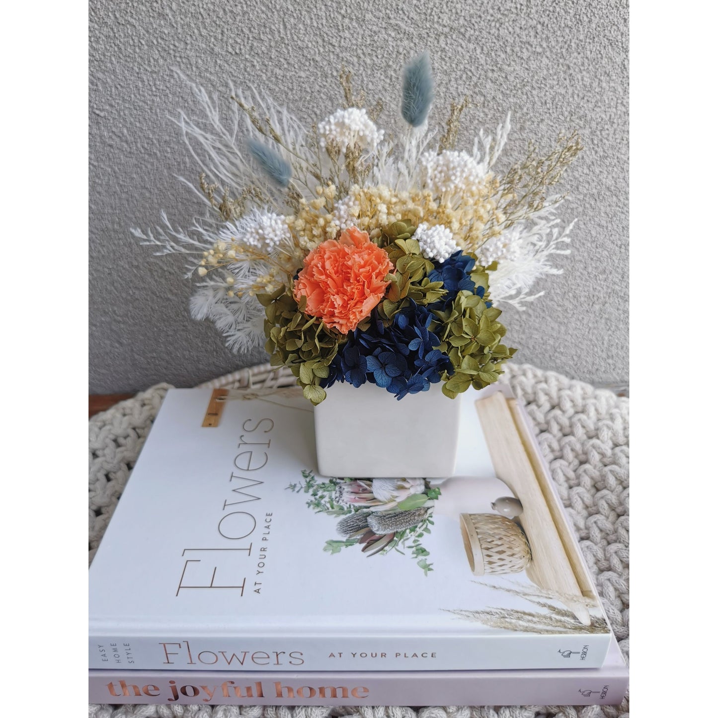 Dried & preserved flower arrangement with navy blue, green, white & orange flowers. Picture shows the arrangement sitting on books on a table in front of a blank wall