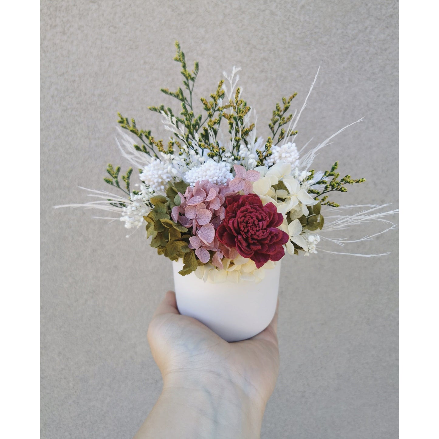 Dried & Preserved flower arrangement with green , white, yellow, pink & burgundy colours. Photo shows arrangement being held by hand in front of a blank wall with a closer view of flowers