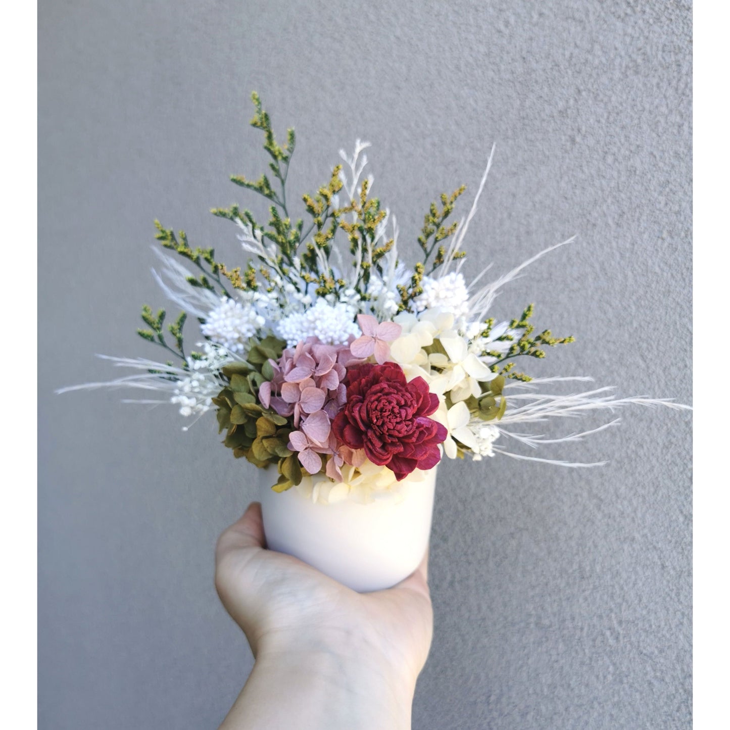 Dried & Preserved flower arrangement with green , white, yellow, pink & burgundy colours. Photo shows arrangement being held by hand in front of a blank wall