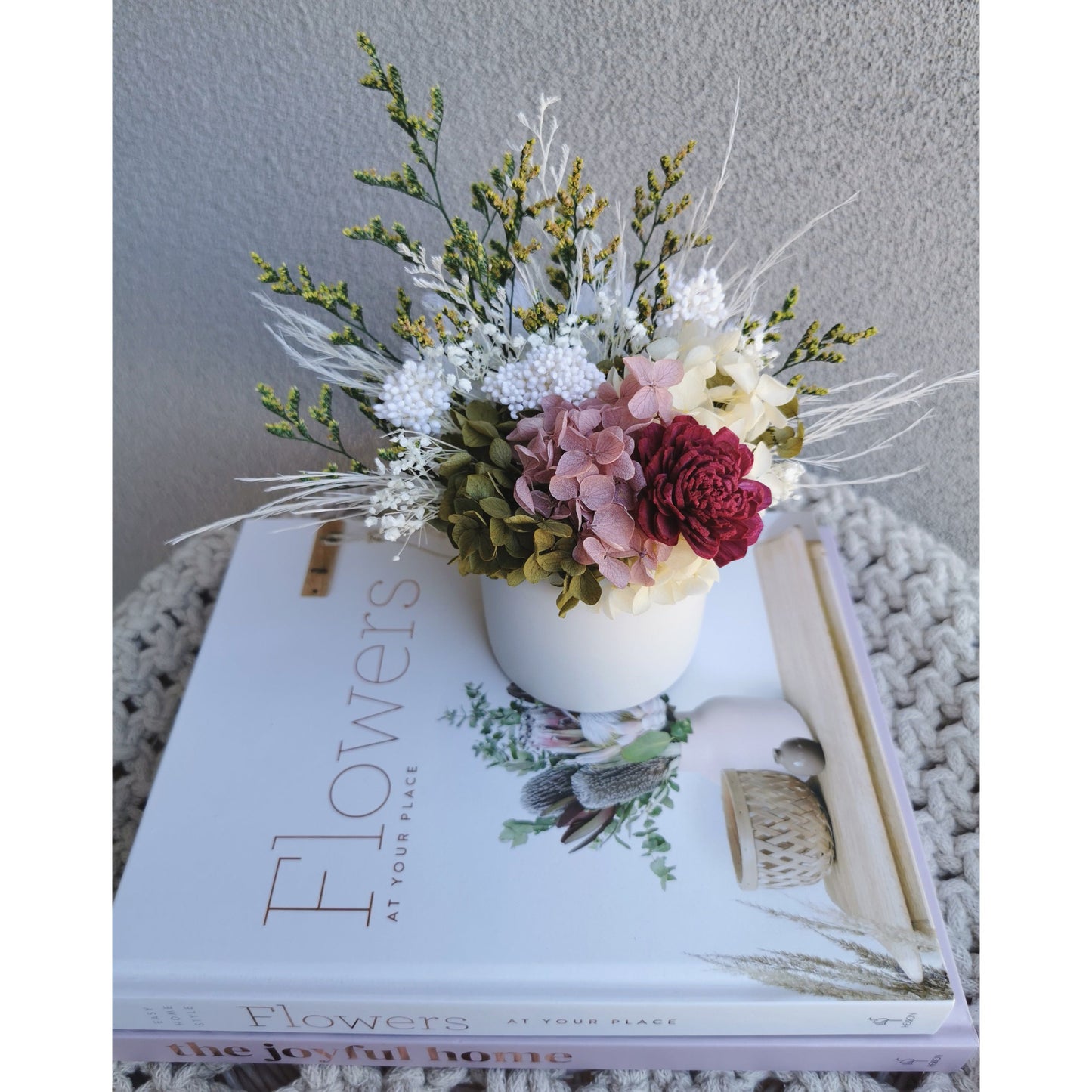 Dried & Preserved flower arrangement with green , white, yellow, pink & burgundy colours. Photo shows arrangement sitting on books in front of a blank wall with a more birds eye view