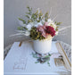 Dried & Preserved flower arrangement with green , white, yellow, pink & burgundy colours. Photo shows arrangement sitting on books in front of a blank wall