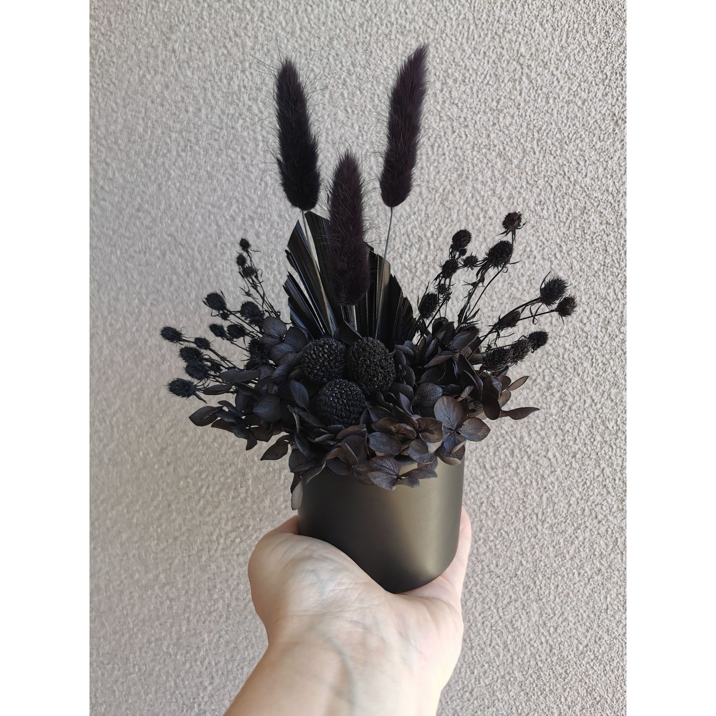 Dried flower arrangement with all black flowers in a black ceramic pot. Photo shows arrangement being held by hand against a blank wall