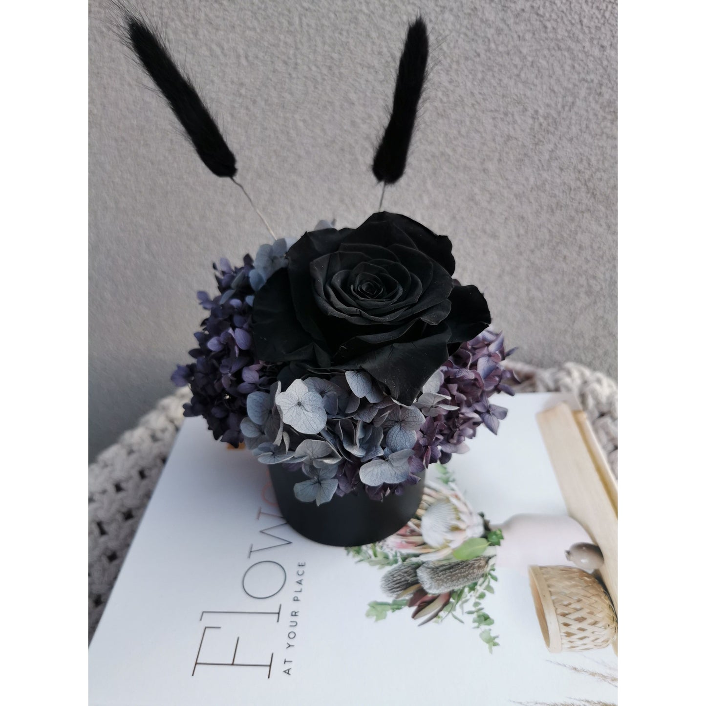 Black, grey & purple dried & preserved flower arrangement in mini black pot featuring a black rose and black bunny tails. Photo shows arrangement sitting on top of books against a blank wall