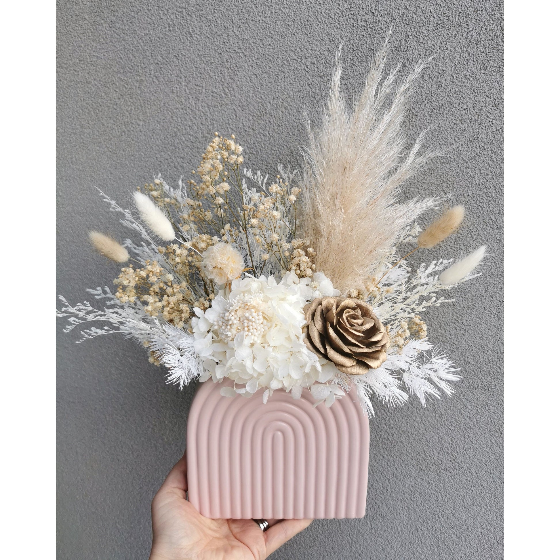 Natural beige and white dried & preserved flower arrangement featuring a gold sola wood flower rose and set in to a pink arch vase. Photo shows arrangement being held by hand against a blank wall