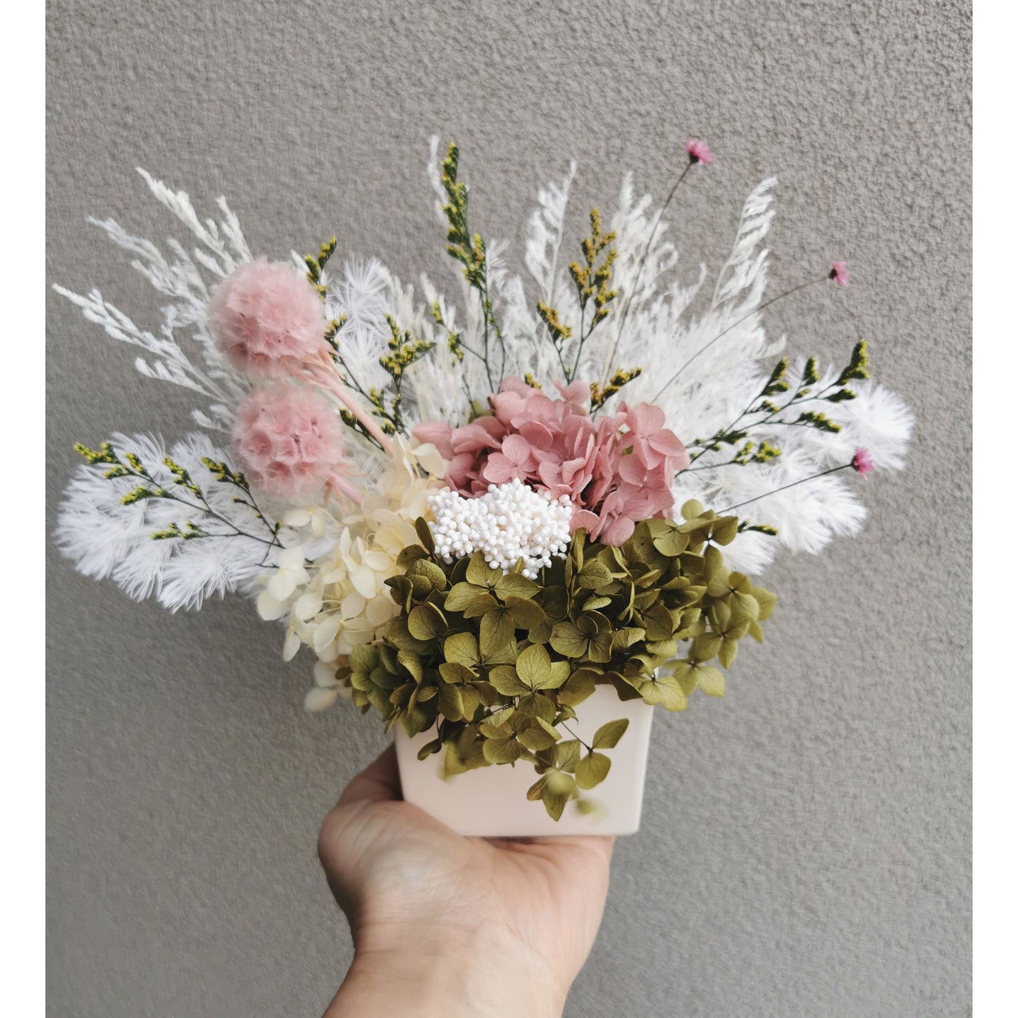 Pink, green and white dried and preserved flower arrangement in white square pot. Photo shows arrangement being held by hand against a blank wall