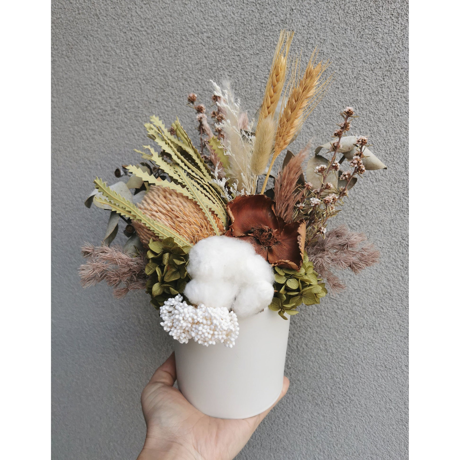 Mix of native dried flowers set in to a round white pot. Photo shows arrangement being held by hand against a blank wall