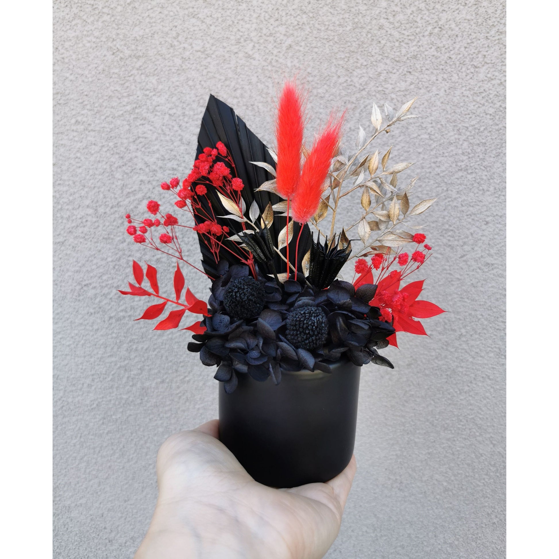 Red, black & gold dried & preserved flowers in black vase. Photo shows arrangement being held by hand against a blank wall