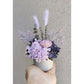 Purple dried & preserved flowers in mini ribbed pot. Photo shows arrangement being held by hand against a blank walll