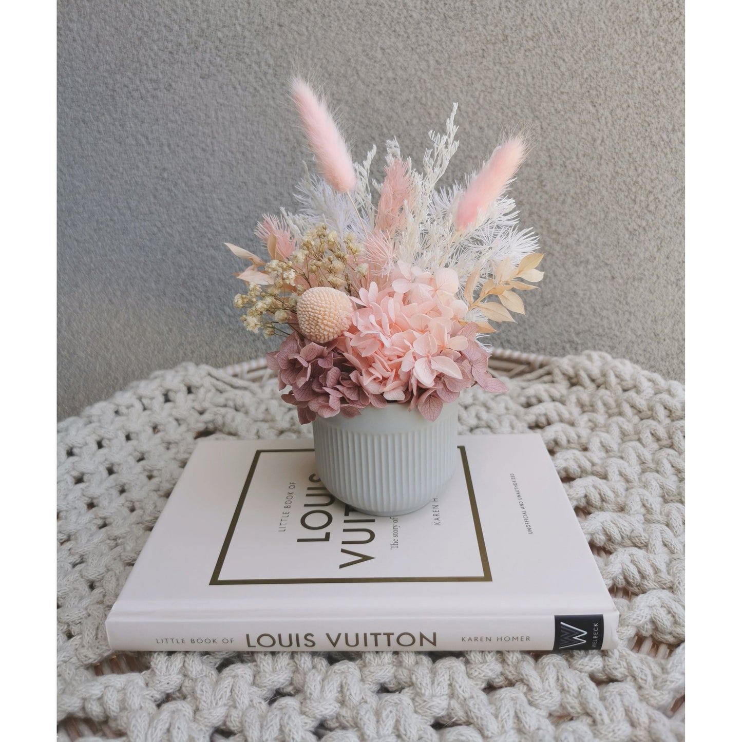 Mini pink dried & preserved flower arrangement in mini ribbed grey pot. Photo shows arrangement sitting on a book against a blank wall
