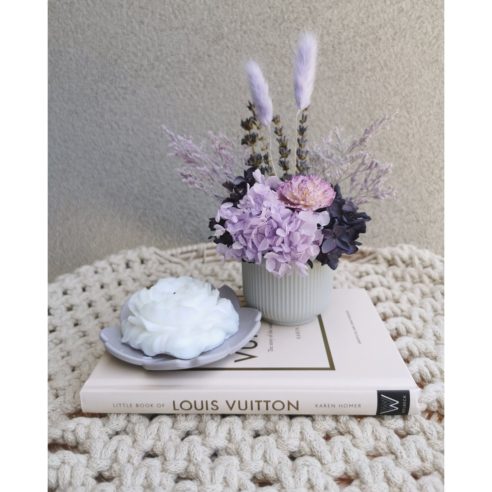 Purple dried & preserved flowers in mini ribbed pot. Photo shows arrangement sitting on a book against a blank wall