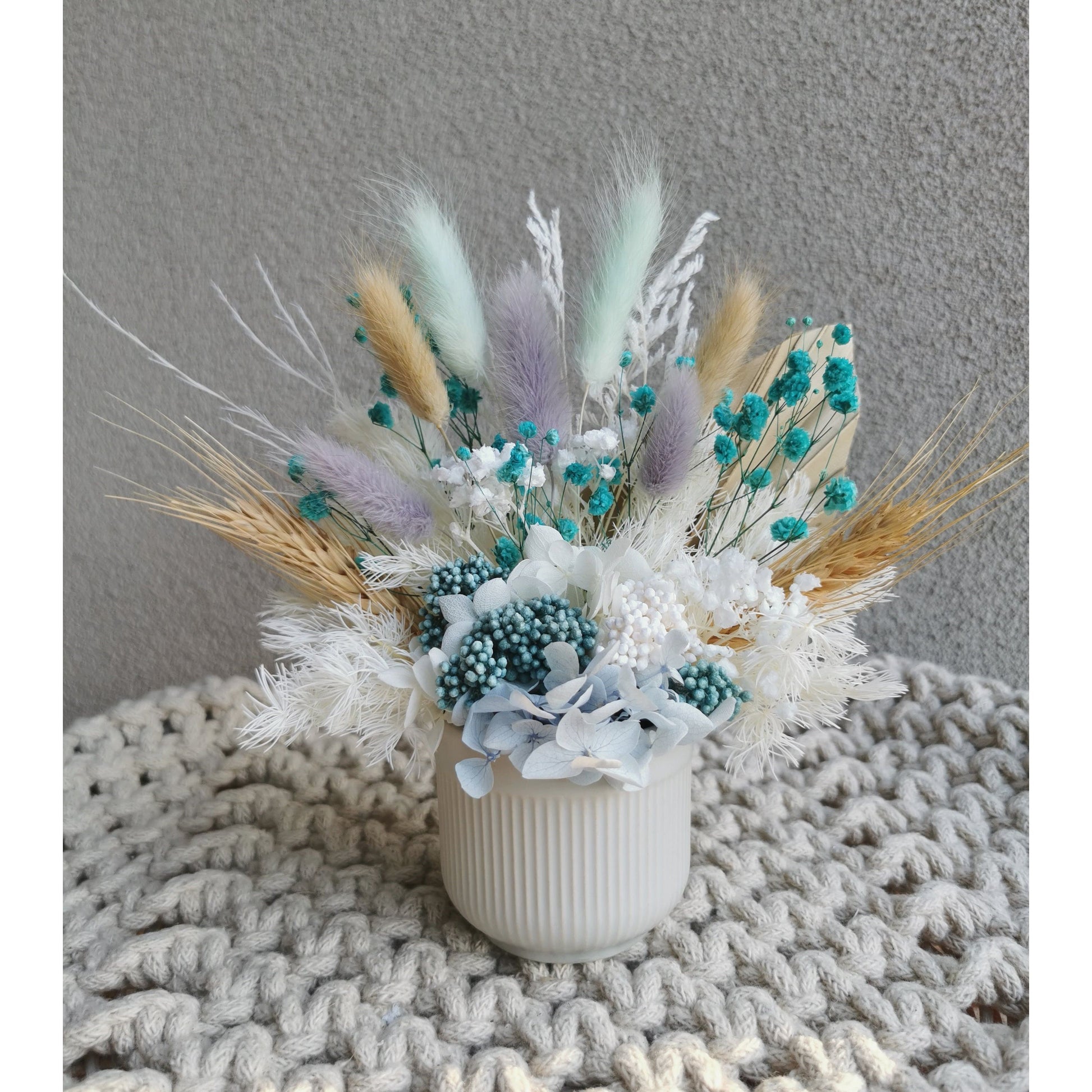 Natural, white, blue & purple dried & preserved mini flower arrangement in ribbed pot. Photo shows arrangement sitting on a tray against a blank wall