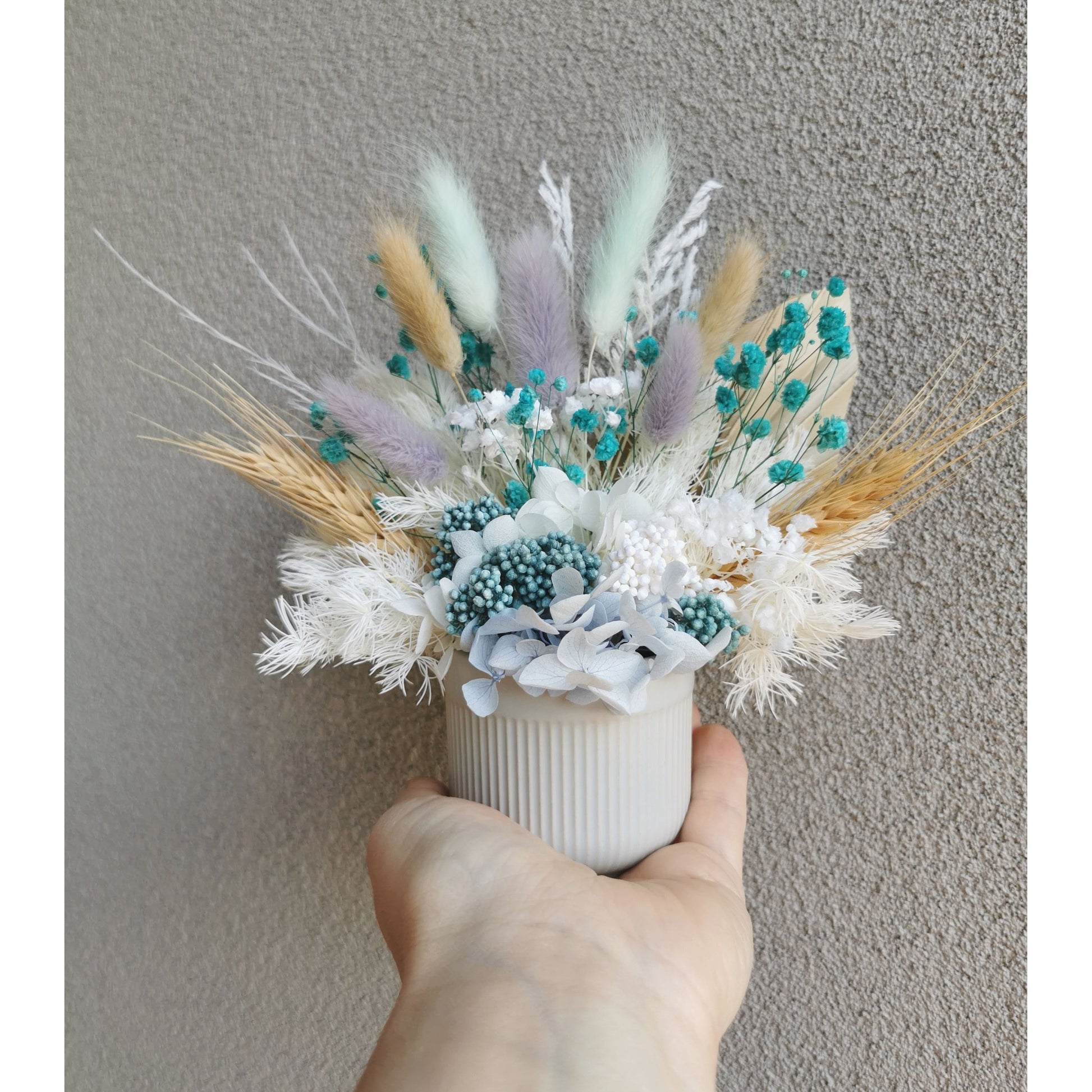 Natural, white, blue & purple dried & preserved mini flower arrangement in ribbed pot. Photo shows arrangement being held by hand against a blank wall