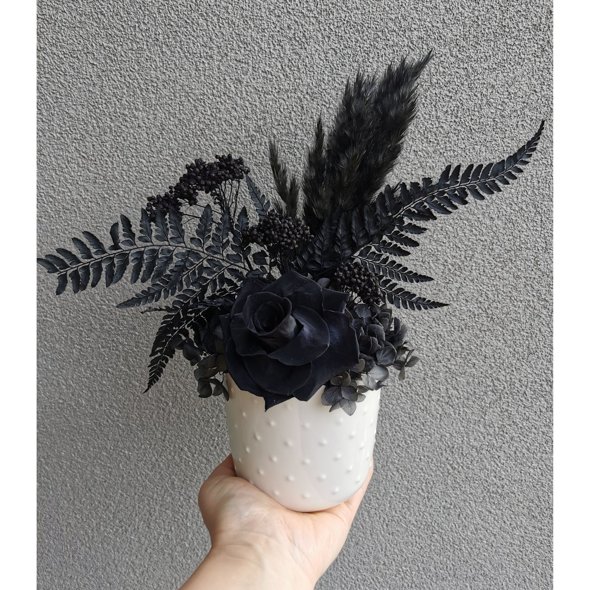 Dried & Preserved flower arrangement featuring all black flowers including a rose, pampas grass, hydrangea, rice flower & ferns. Photo shows arrangement being held by hand against a blank wall