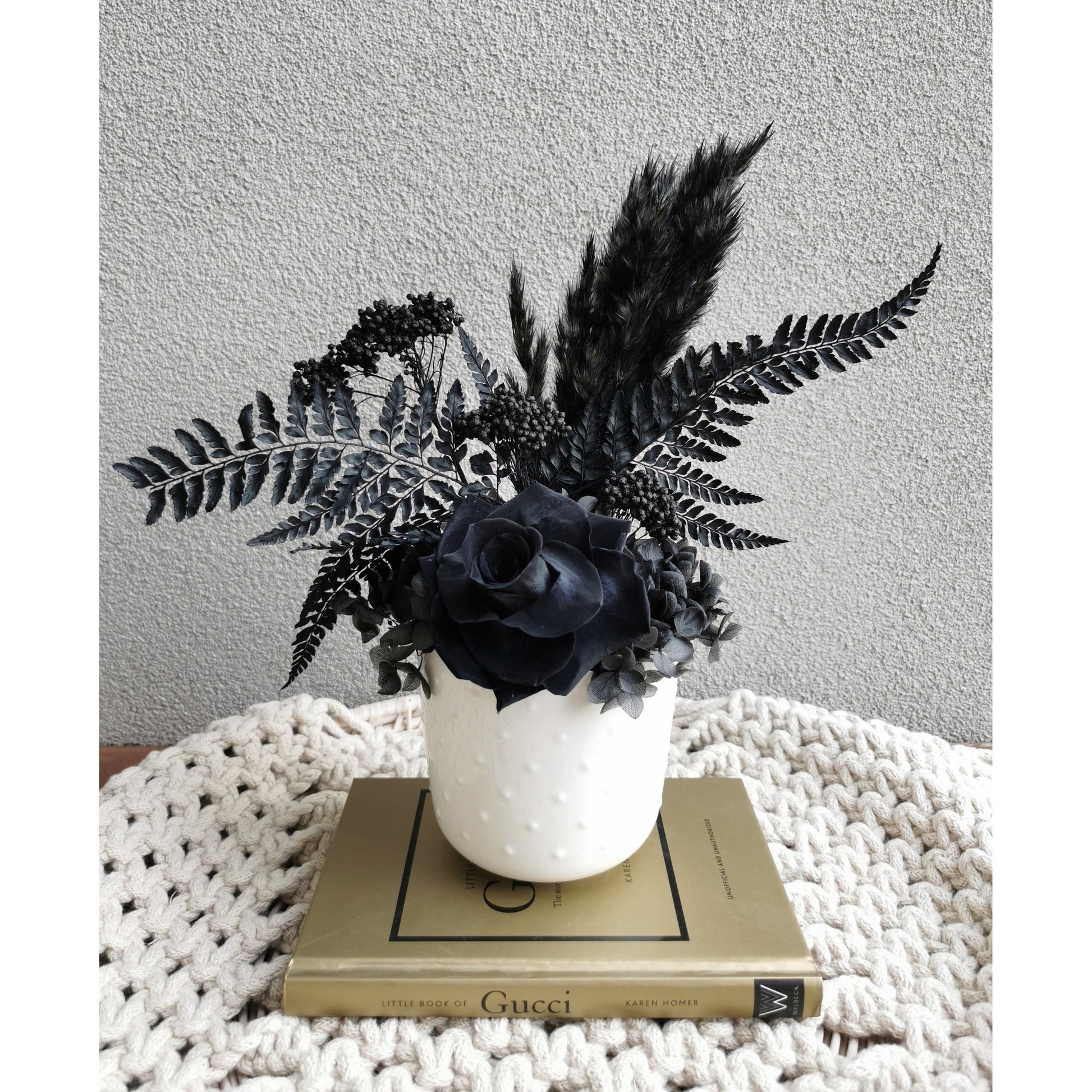 Dried & Preserved flower arrangement featuring all black flowers including a rose, pampas grass, hydrangea, rice flower & ferns. Photo shows arrangement sitting on a book on a table against a blank wall