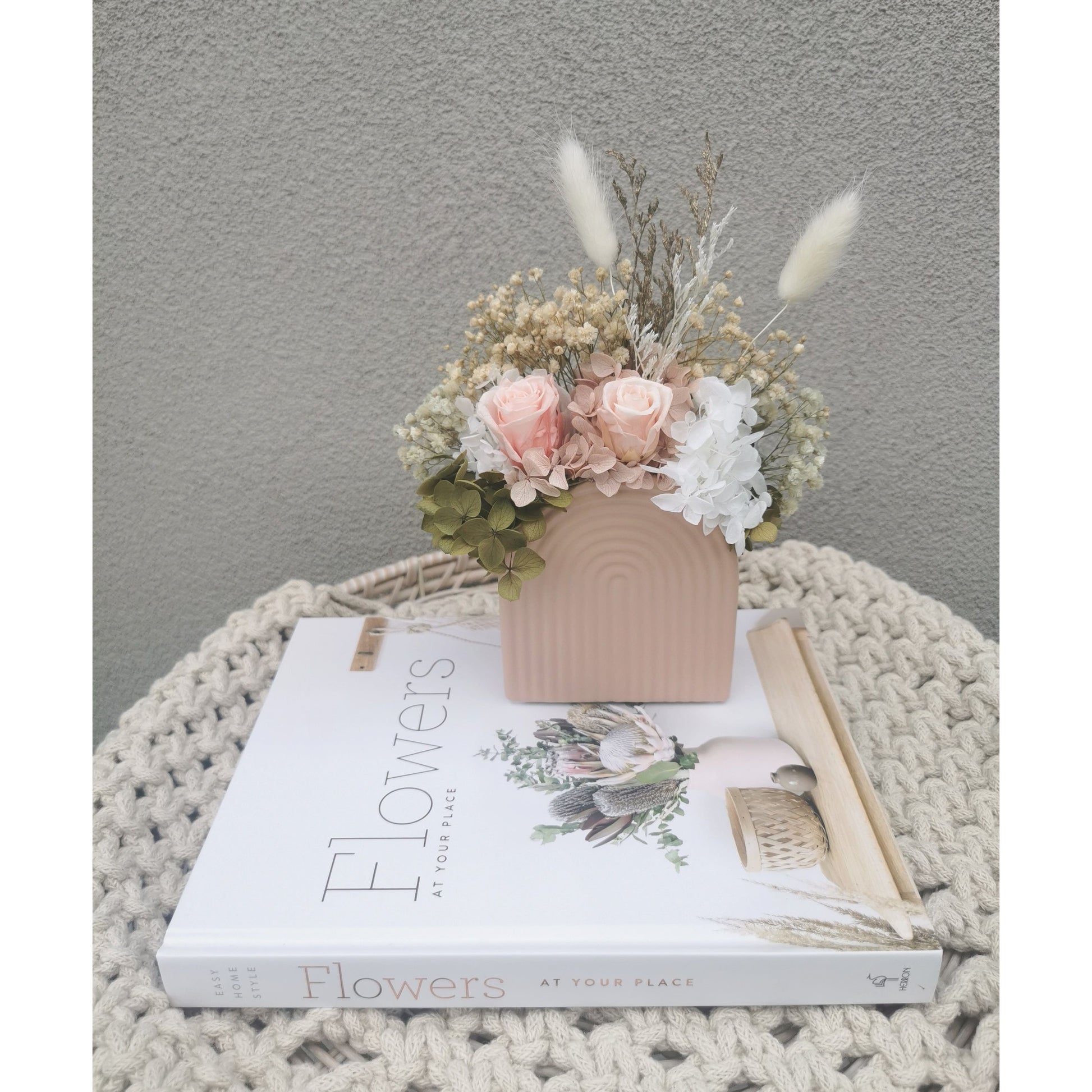 Dried & preserved flower arrangement featuring mini pink roses and pink, green & white dried flowers in a mini pink arch vase. Photo shows arrangement sitting on a book in front of a blank wall
