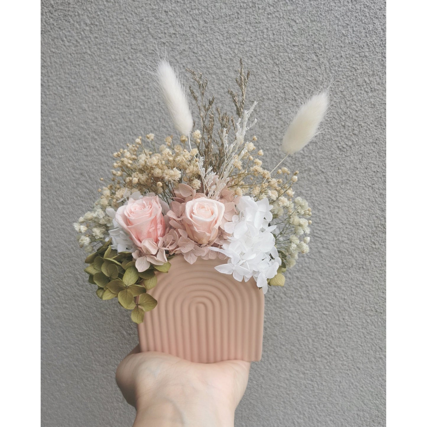 Dried & preserved flower arrangement featuring mini pink roses and pink, green & white dried flowers in a mini pink arch vase. Photo shows arrangement being held by hand in front of a blank wall