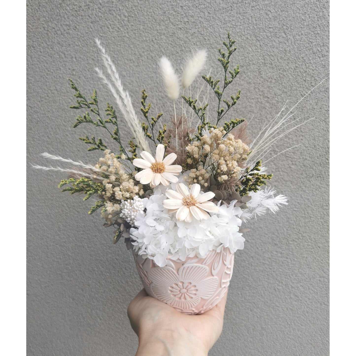 Dried & Preserved flowers in neutral green, white & brown colours in pink & white floral pattern pot. Picture shows arrangement being held by hand against a blank wall