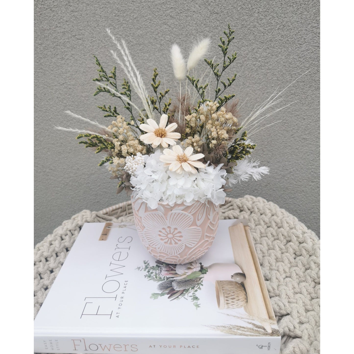 Dried & Preserved flowers in neutral green, white & brown colours in pink & white floral pattern pot. Picture shows arrangement sitting on a book against a blank wall