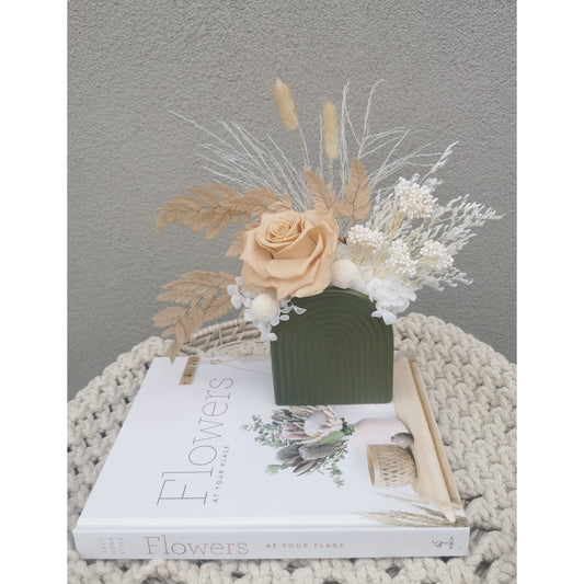 Dried & Preserved flower arrangement with nude and white flowers in a green mini arch vase. Picture shows arragement sitting on a book against a blank wall