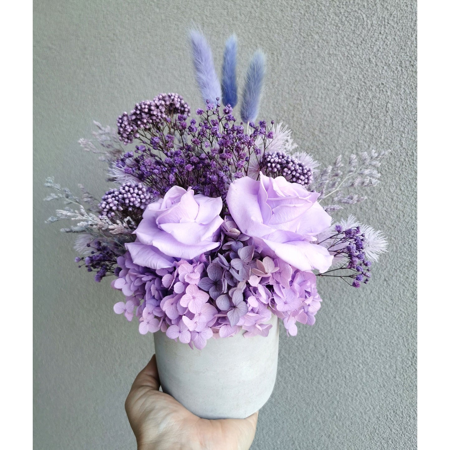 Purple shades of dried & preserved flowers in grey cement pot. Photo shows arrangement being held by hand against a blank wall