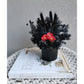 All blacked dried & preserved flowers set in to a mini black pot & featuring 3 mini red flowers in the centre. Photo shows flower arrangement sitting on a book against a blank wall