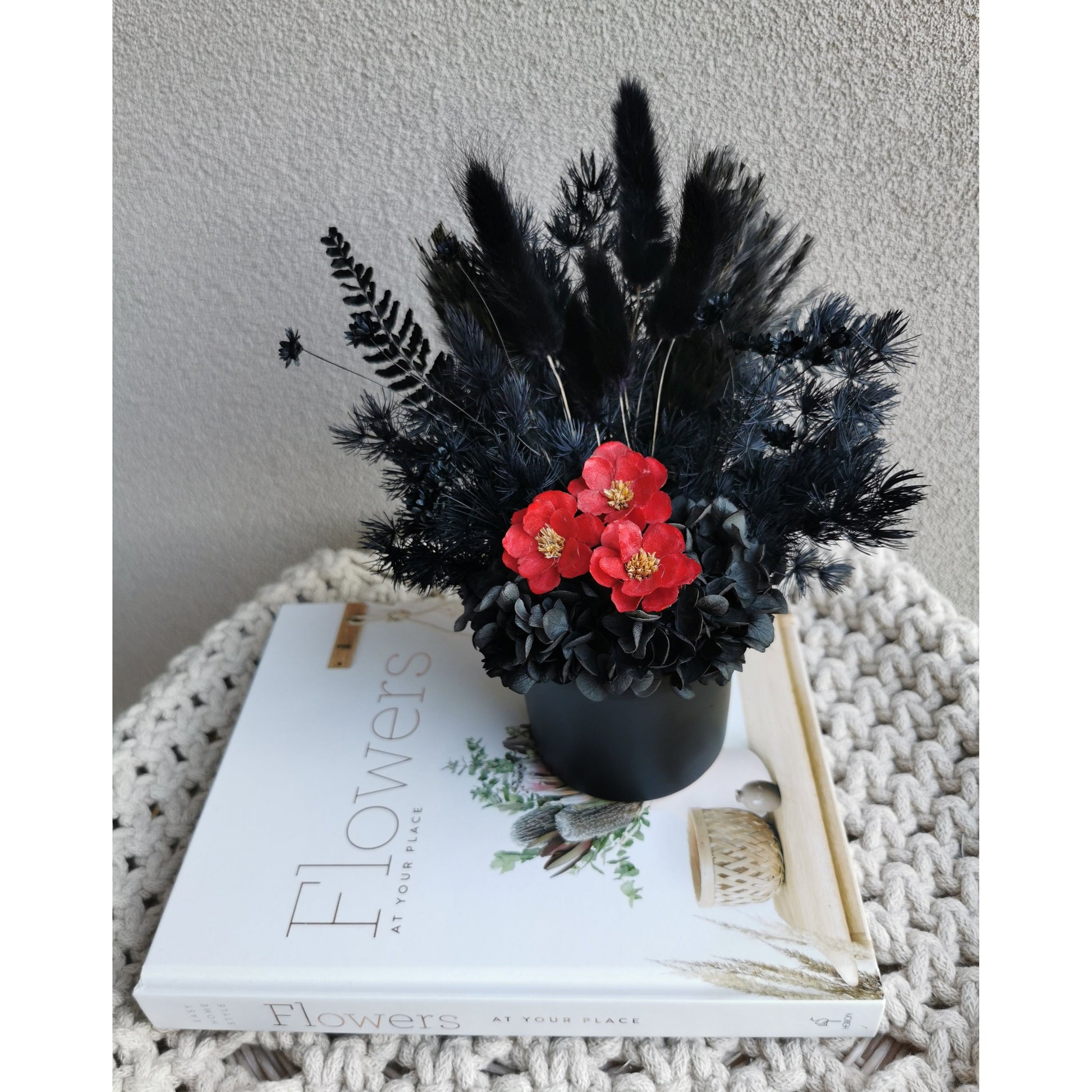 All blacked dried & preserved flowers set in to a mini black pot & featuring 3 mini red flowers in the centre. Photo shows flower arrangement sitting on a book against a blank wall looking down from birds eye view