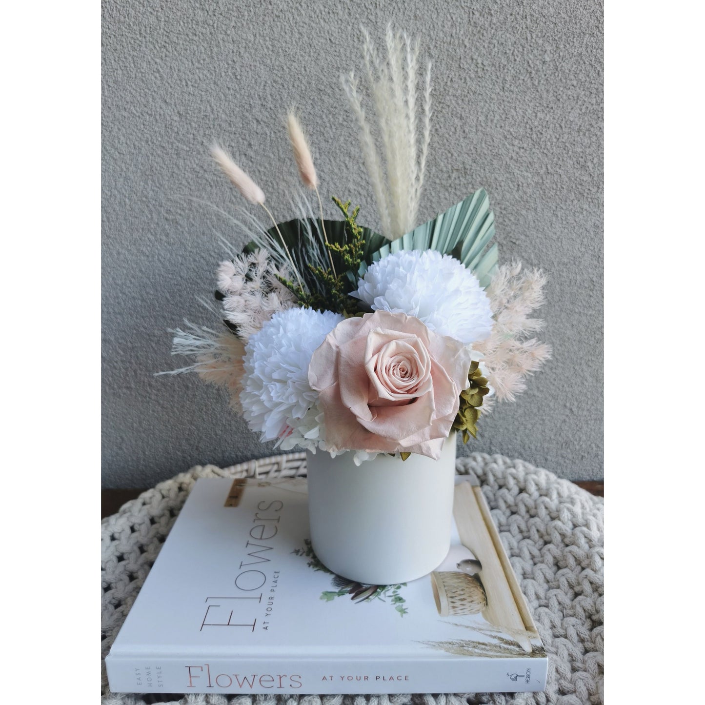 Pink, green & white dried & preserved flower arrangement in white pot. Photo shows arrangement sitting on a book against a blank wall