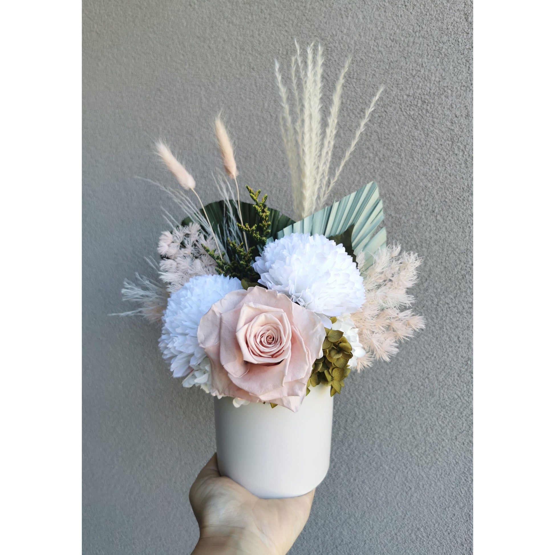 Pink, green & white dried & preserved flower arrangement in white pot. Photo shows arrangement being held by hand against a blank wall