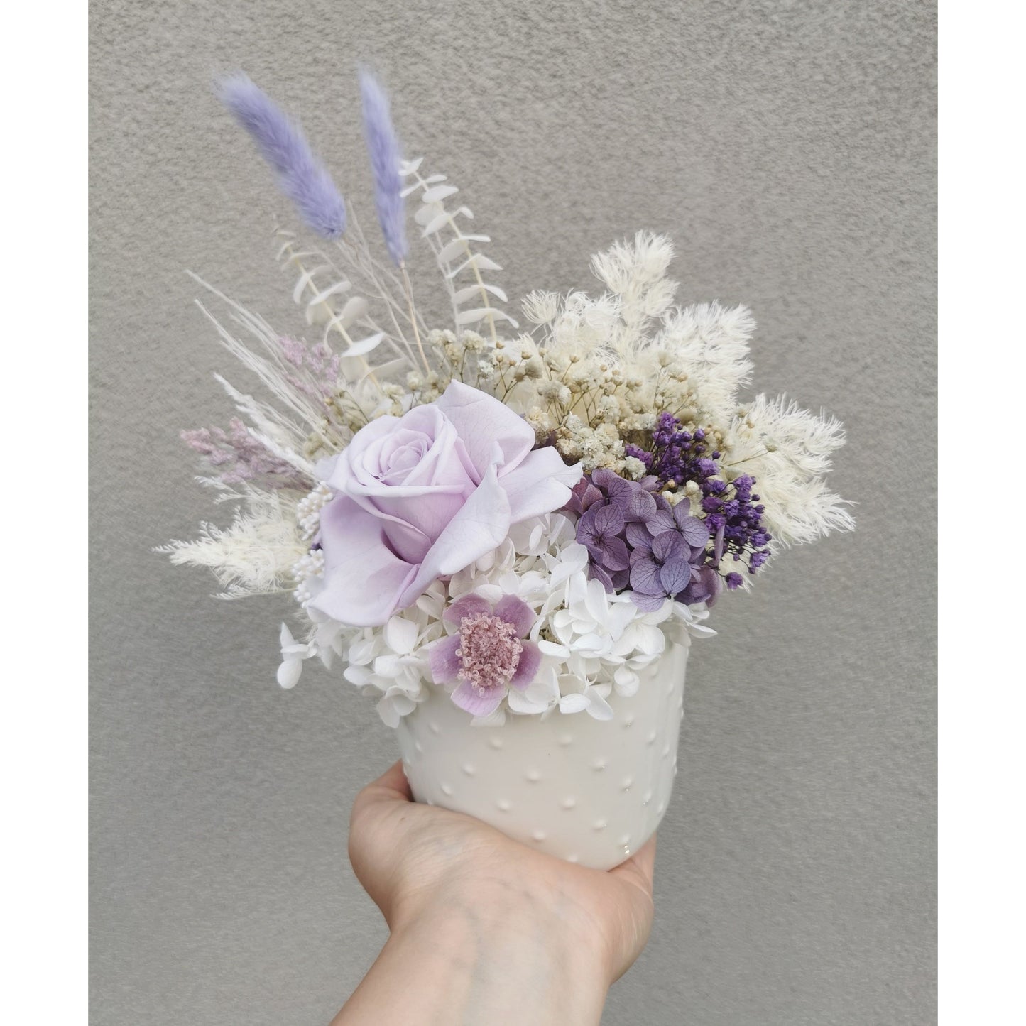 Dried & Preserved flower arrangement in purple & white and featuring a purple preserved rose and nestled in to a white pot. Photo shows arrangement being held by hand against a blank wall