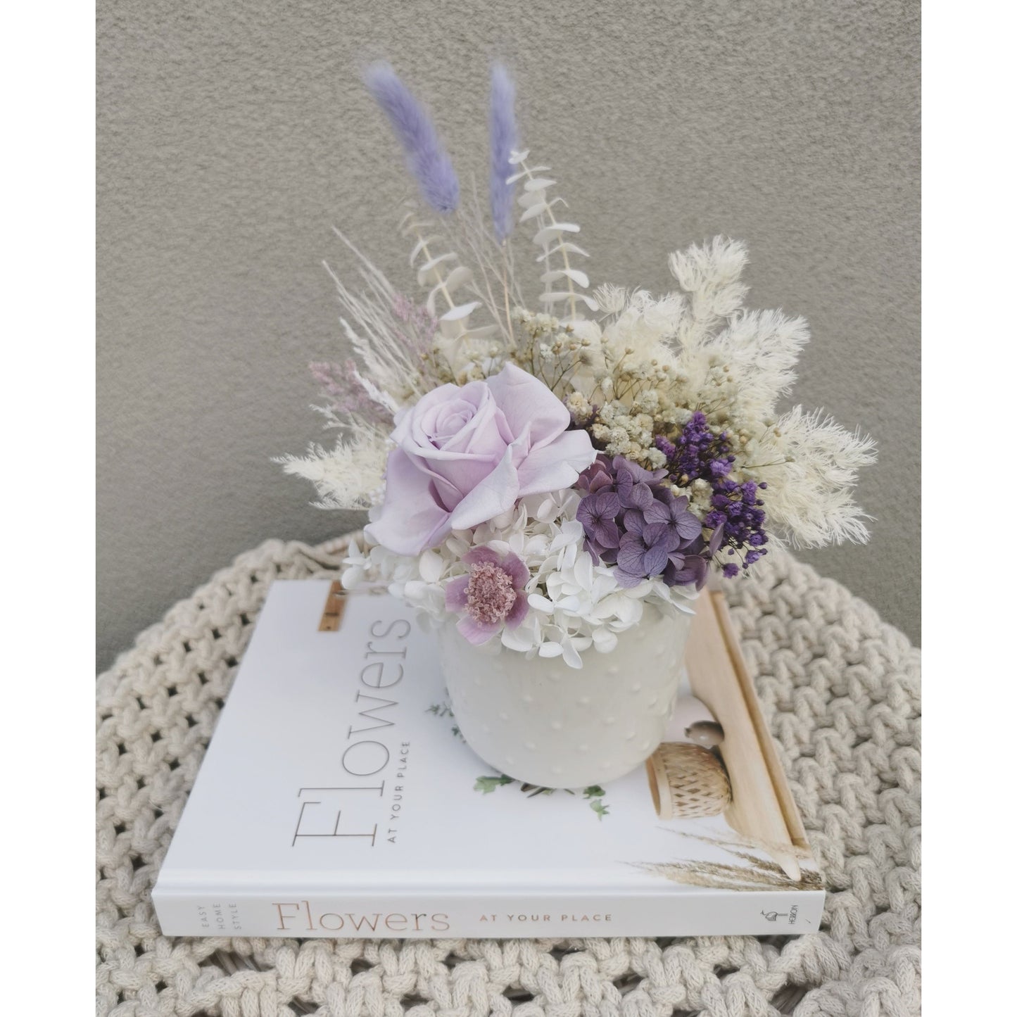 Dried & Preserved flower arrangement in purple & white and featuring a purple preserved rose and nestled in to a white pot. Photo shows arrangement sitting on a book on a table against a blank wall