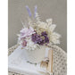 Dried & Preserved flower arrangement in purple & white and featuring a purple preserved rose and nestled in to a white pot. Photo shows arrangement sitting on a left angle view on a book on a table against a blank wall